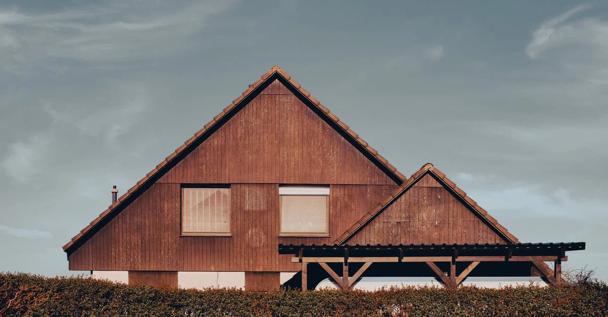 Maison en bois esthétique avec toit triangulaire et arbustes devant, sous un ciel clair.