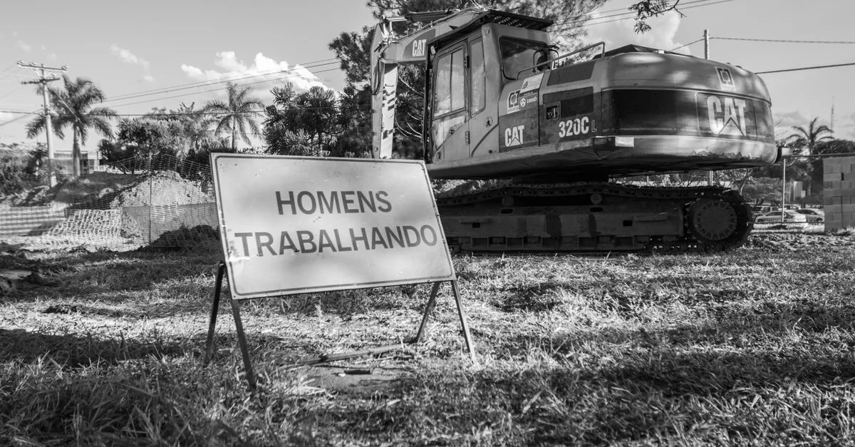 Photo en noir et blanc d'un chantier de construction avec de la machinerie et un panneau en portugais.