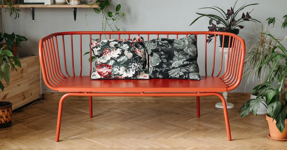 Canapé moderne en métal rouge avec des coussins fleuris entouré de plantes d'intérieur luxuriantes sur un sol en bois.